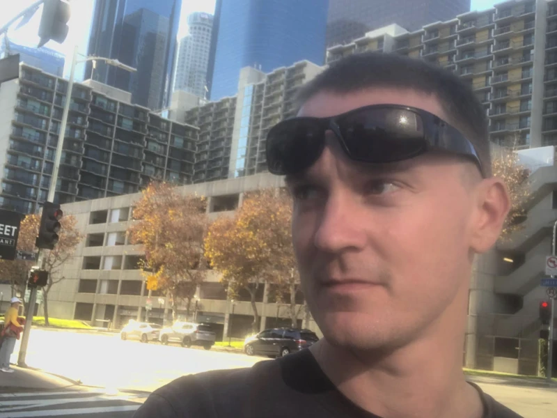 A person with sunglasses on their forehead stands in a city setting. They look off camera.